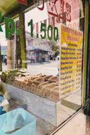 A glass display case contains an assortment of pastries neatly arranged inside. A price list in Indonesian Rupiah is visible on a yellow sign attached to the glass, listing various types of 'Terang Bulan' with different flavor options. The reflection of a street scene can be seen through the glass, including some buildings and a road.