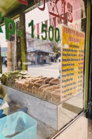 A glass display case contains an assortment of pastries neatly arranged inside. A price list in Indonesian Rupiah is visible on a yellow sign attached to the glass, listing various types of 'Terang Bulan' with different flavor options. The reflection of a street scene can be seen through the glass, including some buildings and a road.