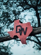 A rusty red metal sign in the shape of Texas is mounted on a wooden beam, with the letters 'DW' prominently attached. The background is a bokeh of tree branches and leaves, creating a textured and atmospheric setting.