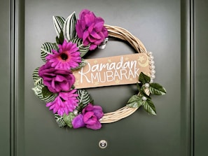 A decorative wreath is displayed on a green door, featuring vibrant purple flowers and green leaves surrounding a beige sign that reads 'Ramadan Mubarak' with star and crescent motifs. Pearls are strung along one side, adding a touch of elegance.
