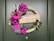 A decorative wreath is displayed on a green door, featuring vibrant purple flowers and green leaves surrounding a beige sign that reads 'Ramadan Mubarak' with star and crescent motifs. Pearls are strung along one side, adding a touch of elegance.