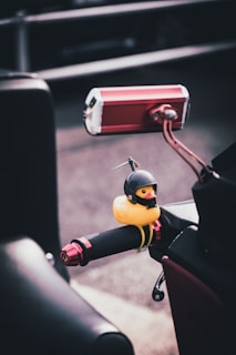 A small, yellow rubber duck dressed in a black helmet is attached to the handle grip of a motorcycle. The grip features a textured surface with red metallic accents. In the background, the road and a blurred section of another motorcycle or vehicle are visible.