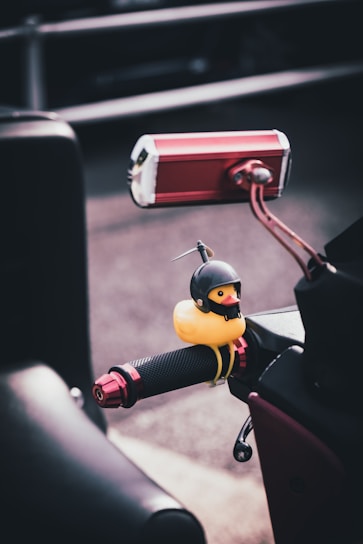A small, yellow rubber duck dressed in a black helmet is attached to the handle grip of a motorcycle. The grip features a textured surface with red metallic accents. In the background, the road and a blurred section of another motorcycle or vehicle are visible.