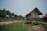 Several rustic wooden huts are raised on stilts in a rural, forested area. The huts have corrugated metal roofs, some of which are slightly rusted. A dirt path winds through the grassy landscape. Dense trees and greenery border the scene, suggesting a secluded, natural environment.