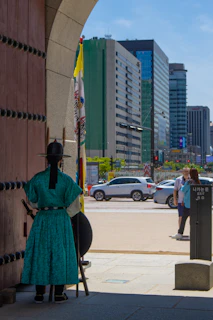 A traditional guard dressed in a teal robe and black hat stands at the entrance of a historic gate, holding a staff and shield. In the background, modern city buildings and busy traffic can be seen under a clear blue sky.