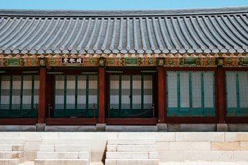 A traditional Korean building features a tiled roof with intricate colorful patterns and wooden framework. The structure has symmetrically arranged doors with latticed windows, situated above stone steps.