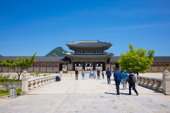 A traditional Korean palace with intricate architectural details stands majestically against a clear blue sky. A group of visitors walks towards the entrance, which is flanked by well-maintained gardens and stone railings. A forested mountain rises in the background, adding to the serene atmosphere.