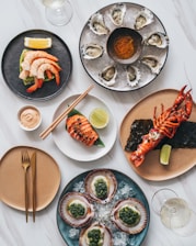 An elegant seafood spread is arranged on a white marble surface. The dishes include oysters on ice with a small bowl of spices or sauce in the center, shrimp on a black plate with avocado and lemon wedge, grilled salmon garnished with herbs and a slice of lime, scallops with green sauce, and a lobster partially split open, accompanied by lime. A glass of white wine is partially visible and there is a neat arrangement of cutlery on one of the plates.