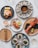 An elegant seafood spread is arranged on a white marble surface. The dishes include oysters on ice with a small bowl of spices or sauce in the center, shrimp on a black plate with avocado and lemon wedge, grilled salmon garnished with herbs and a slice of lime, scallops with green sauce, and a lobster partially split open, accompanied by lime. A glass of white wine is partially visible and there is a neat arrangement of cutlery on one of the plates.