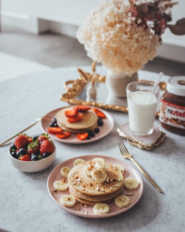 A beautifully arranged breakfast setting on a marble table featuring two plates of pancakes topped with banana slices, strawberries, and nuts. A small bowl filled with fresh strawberries and blueberries is nearby. There is a glass of milk and a jar of Nutella on a small ornate tray. In the background, a bouquet of fluffy white flowers adds a touch of elegance.