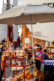 A vibrant street food stall with a large umbrella offers an array of snacks including popcorn, fruit, and various packaged treats. The vendor, wearing a colorful shirt and visor, tends to customers surrounded by an assortment of goods. Nearby signage displays directions and advertisements for minerals and fossils.