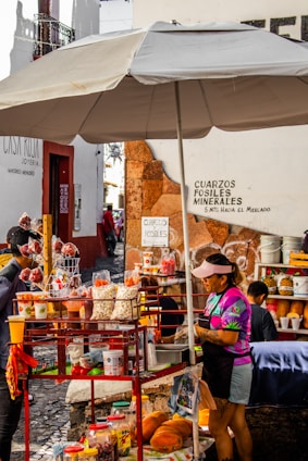 A vibrant street food stall with a large umbrella offers an array of snacks including popcorn, fruit, and various packaged treats. The vendor, wearing a colorful shirt and visor, tends to customers surrounded by an assortment of goods. Nearby signage displays directions and advertisements for minerals and fossils.