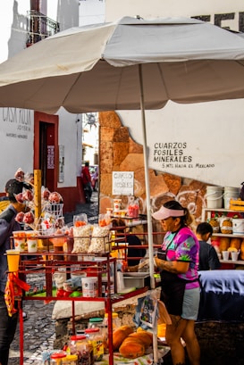 A vibrant street food stall with a large umbrella offers an array of snacks including popcorn, fruit, and various packaged treats. The vendor, wearing a colorful shirt and visor, tends to customers surrounded by an assortment of goods. Nearby signage displays directions and advertisements for minerals and fossils.