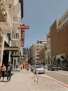 A city street lined with tall buildings, featuring a mix of modern and older architecture styles. The street is bustling with pedestrians walking along the sidewalks, some shopping at high-end stores like Chanel and Jimmy Choo, indicated by their signs. Cars are parked along the street, and some are driving past. A prominent red sign for a fabric store adds a splash of color to the scene.