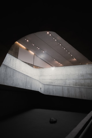 A minimalist architectural space with concrete walls and a high ceiling. The lighting is provided by several small, evenly spaced lights on the ceiling, casting a subtle glow over the area. The walls form a modern, angular design with a light gray color. At the bottom, a dark object is resting on a dark floor.