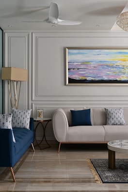 A sophisticated living room with elegant furniture and decor. A cream-colored couch is adorned with blue accent pillows, complemented by a navy-blue sofa featuring patterned cushions. Between the sofas, there is a small round side table with a decorative item on top. The floor is made of polished stone tiles and a patterned rug partially covers it. The walls are decorated with paneling and a large abstract painting depicting a colorful seascape. A ceiling fan and a floor lamp add to the room's design.