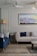 A sophisticated living room with elegant furniture and decor. A cream-colored couch is adorned with blue accent pillows, complemented by a navy-blue sofa featuring patterned cushions. Between the sofas, there is a small round side table with a decorative item on top. The floor is made of polished stone tiles and a patterned rug partially covers it. The walls are decorated with paneling and a large abstract painting depicting a colorful seascape. A ceiling fan and a floor lamp add to the room's design.