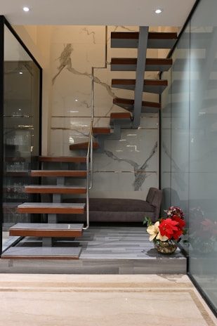 A modern interior featuring a floating wooden staircase with metal supports, set against a wall with marble-like patterns. The floor is lined with light gray wood, and beside the staircase is a large glass wall. A plush brown bench is placed at the corner alongside a decorative vase containing red and cream-colored flowers.