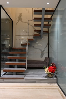 A modern interior featuring a floating wooden staircase with metal supports, set against a wall with marble-like patterns. The floor is lined with light gray wood, and beside the staircase is a large glass wall. A plush brown bench is placed at the corner alongside a decorative vase containing red and cream-colored flowers.