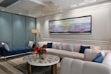 A well-furnished living room with modern decor. The room features a large, U-shaped white sofa with blue and white patterned pillows, a blue bench, and a round coffee table with a decorative black and white pattern. Red flowers in a vase sit on the coffee table. A large, abstract painting with hues of yellow, purple, and blue hangs on the wall behind the sofa. A chandelier hangs from the ceiling, and a tall floor lamp is placed near the blue bench. The floor is a combination of light and dark marble tiles, and there are glass partitions in the background.