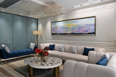 A well-furnished living room with modern decor. The room features a large, U-shaped white sofa with blue and white patterned pillows, a blue bench, and a round coffee table with a decorative black and white pattern. Red flowers in a vase sit on the coffee table. A large, abstract painting with hues of yellow, purple, and blue hangs on the wall behind the sofa. A chandelier hangs from the ceiling, and a tall floor lamp is placed near the blue bench. The floor is a combination of light and dark marble tiles, and there are glass partitions in the background.