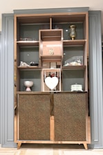 A wooden cabinet with several shelves contains decorative items, including a golden vase, a small sculpture, a heart-shaped ornament, and a variety of other decorative pieces. The cabinet has a patterned design on its lower half, adding a touch of elegance. The shelving units are framed by a stylish border.