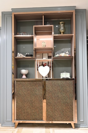 A wooden cabinet with several shelves contains decorative items, including a golden vase, a small sculpture, a heart-shaped ornament, and a variety of other decorative pieces. The cabinet has a patterned design on its lower half, adding a touch of elegance. The shelving units are framed by a stylish border.