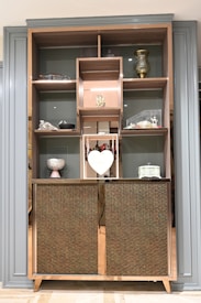 A wooden cabinet with several shelves contains decorative items, including a golden vase, a small sculpture, a heart-shaped ornament, and a variety of other decorative pieces. The cabinet has a patterned design on its lower half, adding a touch of elegance. The shelving units are framed by a stylish border.