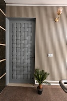 A modern interior door with a woven pattern design positioned next to a ribbed beige wall. There is a small potted plant at the base of the wall and a light fixture with a decorative bulb mounted above. The area has a carpeted floor and a light switch is visible on the wall.