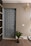 A modern interior door with a woven pattern design positioned next to a ribbed beige wall. There is a small potted plant at the base of the wall and a light fixture with a decorative bulb mounted above. The area has a carpeted floor and a light switch is visible on the wall.