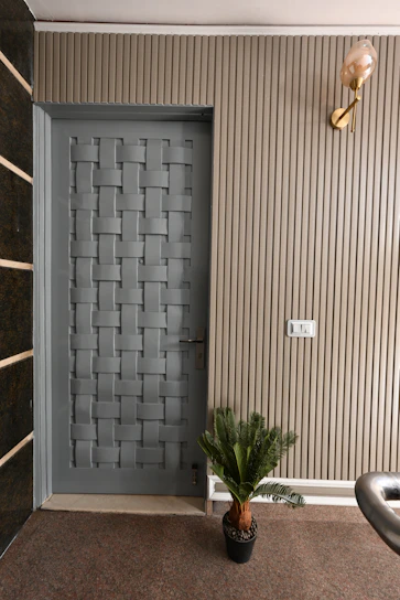 A modern interior door with a woven pattern design positioned next to a ribbed beige wall. There is a small potted plant at the base of the wall and a light fixture with a decorative bulb mounted above. The area has a carpeted floor and a light switch is visible on the wall.