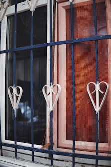 A metal window grill with heart-shaped decorations is prominently featured. The grill is painted in a dark blue color. Behind the grill, there is a window with white framing and a reddish-orange mesh protecting it. The materials appear weathered, adding a rustic feeling to the scene.