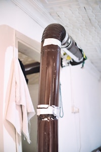A large, brown metal pipe is installed along the corner of a room. The pipe is supported by several bands of white tape or insulation material. Adjacent to the pipe, a white cloth or towel is hanging on a hook. The walls and ceiling are white, with a textured ceiling pattern, and there are visible cables or wires near the pipe.