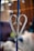 A metal fence decoration, shaped like a heart, is attached to a vertical bar. The decoration appears to be weathered with some rust. The background is blurred, featuring warm-toned colors and indistinct shapes, suggesting an indoor or sheltered setting.
