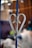 A metal fence decoration, shaped like a heart, is attached to a vertical bar. The decoration appears to be weathered with some rust. The background is blurred, featuring warm-toned colors and indistinct shapes, suggesting an indoor or sheltered setting.