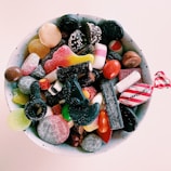 A ceramic bowl filled with a variety of colorful candies, including gummy sweets, licorice, chocolates, and sugar-coated pieces. The candies display a mix of textures, from shiny to matte and sugary surfaces.