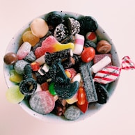A ceramic bowl filled with a variety of colorful candies, including gummy sweets, licorice, chocolates, and sugar-coated pieces. The candies display a mix of textures, from shiny to matte and sugary surfaces.