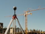 A tripod-mounted surveying instrument is prominently placed in the foreground, with a large construction site visible in the background. Tall cranes and unfinished buildings indicate ongoing construction work, set against a clear blue sky.