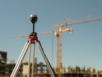 A tripod-mounted surveying instrument is prominently placed in the foreground, with a large construction site visible in the background. Tall cranes and unfinished buildings indicate ongoing construction work, set against a clear blue sky.