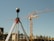 A tripod-mounted surveying instrument is prominently placed in the foreground, with a large construction site visible in the background. Tall cranes and unfinished buildings indicate ongoing construction work, set against a clear blue sky.