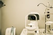 A white optometry machine is situated on an examination table, used for eye testing, alongside other medical equipment in a clinical setting. The background is neutral with smooth walls.