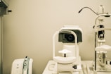 A white optometry machine is situated on an examination table, used for eye testing, alongside other medical equipment in a clinical setting. The background is neutral with smooth walls.