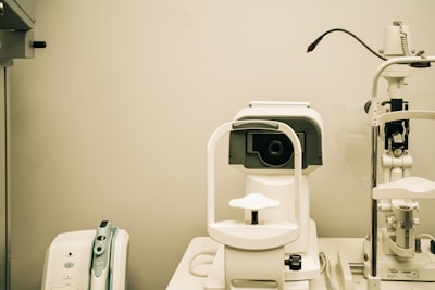 A white optometry machine is situated on an examination table, used for eye testing, alongside other medical equipment in a clinical setting. The background is neutral with smooth walls.