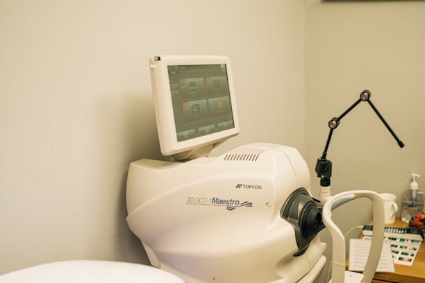 A medical imaging device with a monitor displaying a user interface featuring multiple icons and buttons. The device is labeled 3D OCT Maestro by Topcon. There is also a black adjustable arm and other medical supplies in the background.