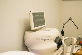 A medical imaging device with a monitor displaying a user interface featuring multiple icons and buttons. The device is labeled 3D OCT Maestro by Topcon. There is also a black adjustable arm and other medical supplies in the background.