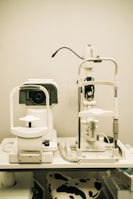 A pair of optical instruments sit on a table, commonly found in an optometrist's office. The setup includes a phoropter and a slit lamp, both primarily white with some black and metallic components. A focus knob is visible on the side of one of the devices, and there's a cable connected to the other. The background is plain, highlighting the clinical environment.