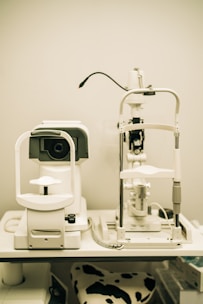 A pair of optical instruments sit on a table, commonly found in an optometrist's office. The setup includes a phoropter and a slit lamp, both primarily white with some black and metallic components. A focus knob is visible on the side of one of the devices, and there's a cable connected to the other. The background is plain, highlighting the clinical environment.