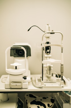 A pair of optical instruments sit on a table, commonly found in an optometrist's office. The setup includes a phoropter and a slit lamp, both primarily white with some black and metallic components. A focus knob is visible on the side of one of the devices, and there's a cable connected to the other. The background is plain, highlighting the clinical environment.