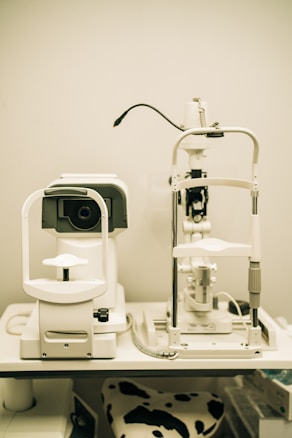 A pair of optical instruments sit on a table, commonly found in an optometrist's office. The setup includes a phoropter and a slit lamp, both primarily white with some black and metallic components. A focus knob is visible on the side of one of the devices, and there's a cable connected to the other. The background is plain, highlighting the clinical environment.