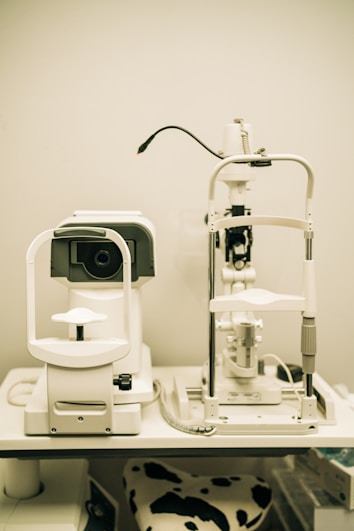 A pair of optical instruments sit on a table, commonly found in an optometrist's office. The setup includes a phoropter and a slit lamp, both primarily white with some black and metallic components. A focus knob is visible on the side of one of the devices, and there's a cable connected to the other. The background is plain, highlighting the clinical environment.